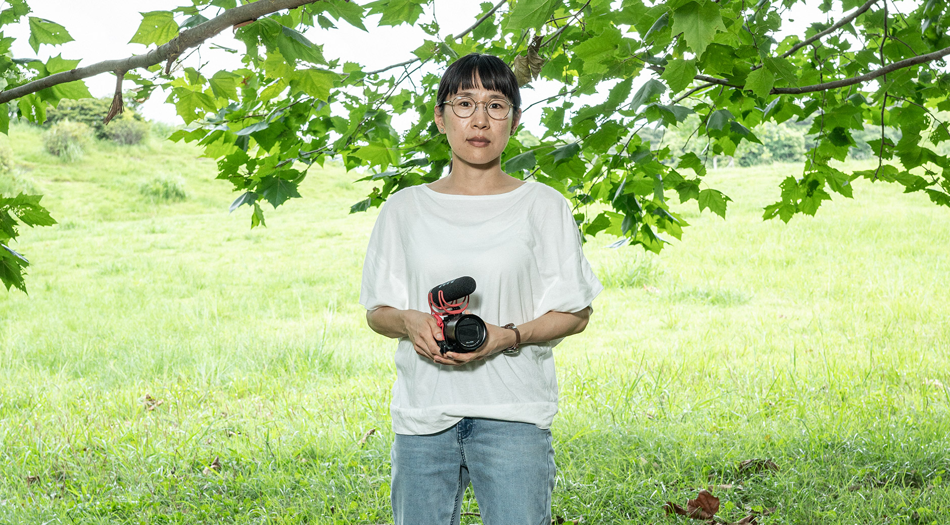 【画像】ビデオカメラをもち、木の下に立ついまむらあやこさん