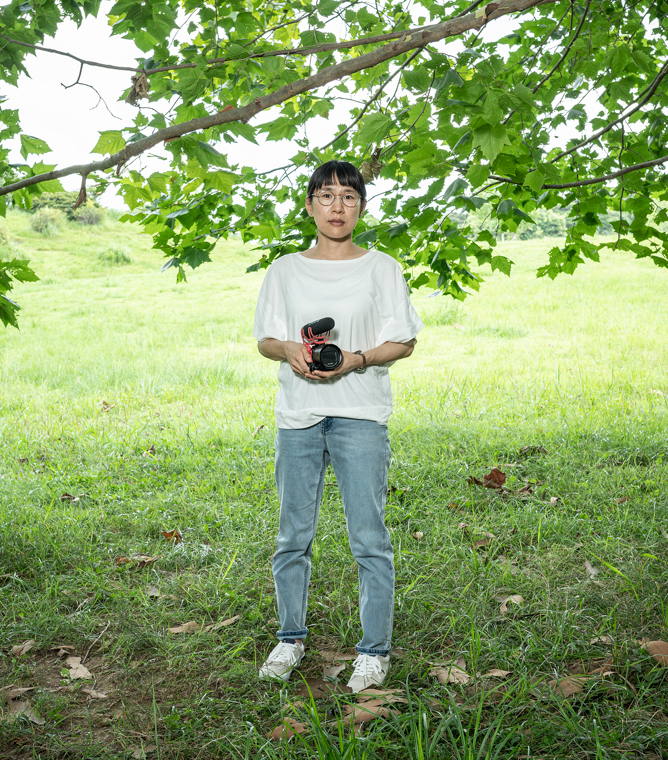 【画像】ビデオカメラをもち、木の下に立ついまむらあやこさん