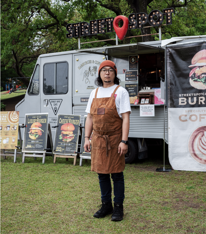 【写真】キッチンカーの前に立つ吉田茂樹さん。オレンジ色のニットキャップにメガネ、長髪、ヒゲ、茶色いエプロンをつけている。
