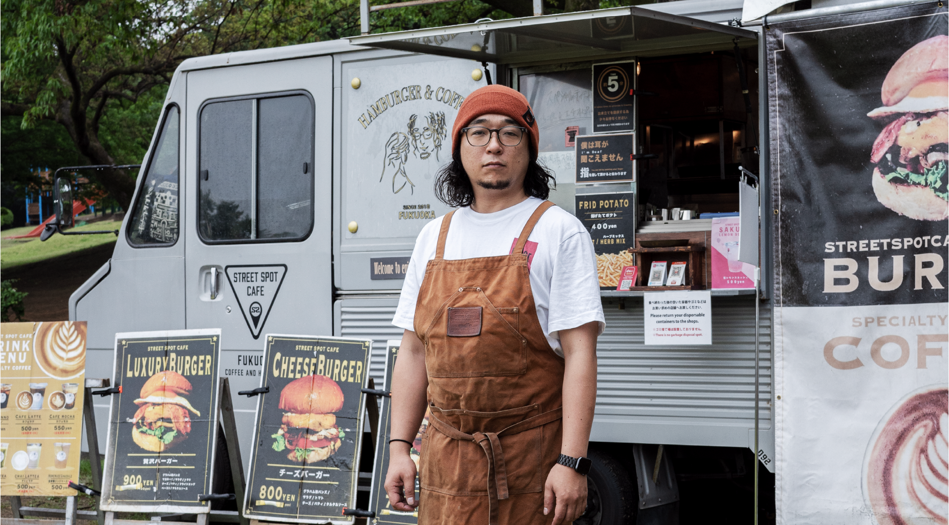 【写真】キッチンカーの前に立つ吉田茂樹さん。オレンジ色のニットキャップにメガネ、長髪、ヒゲ、茶色いエプロンをつけている。