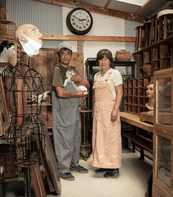 【写真】古道具が並ぶ倉庫の中で、妻と猫とともに佇む 西川さとしさん