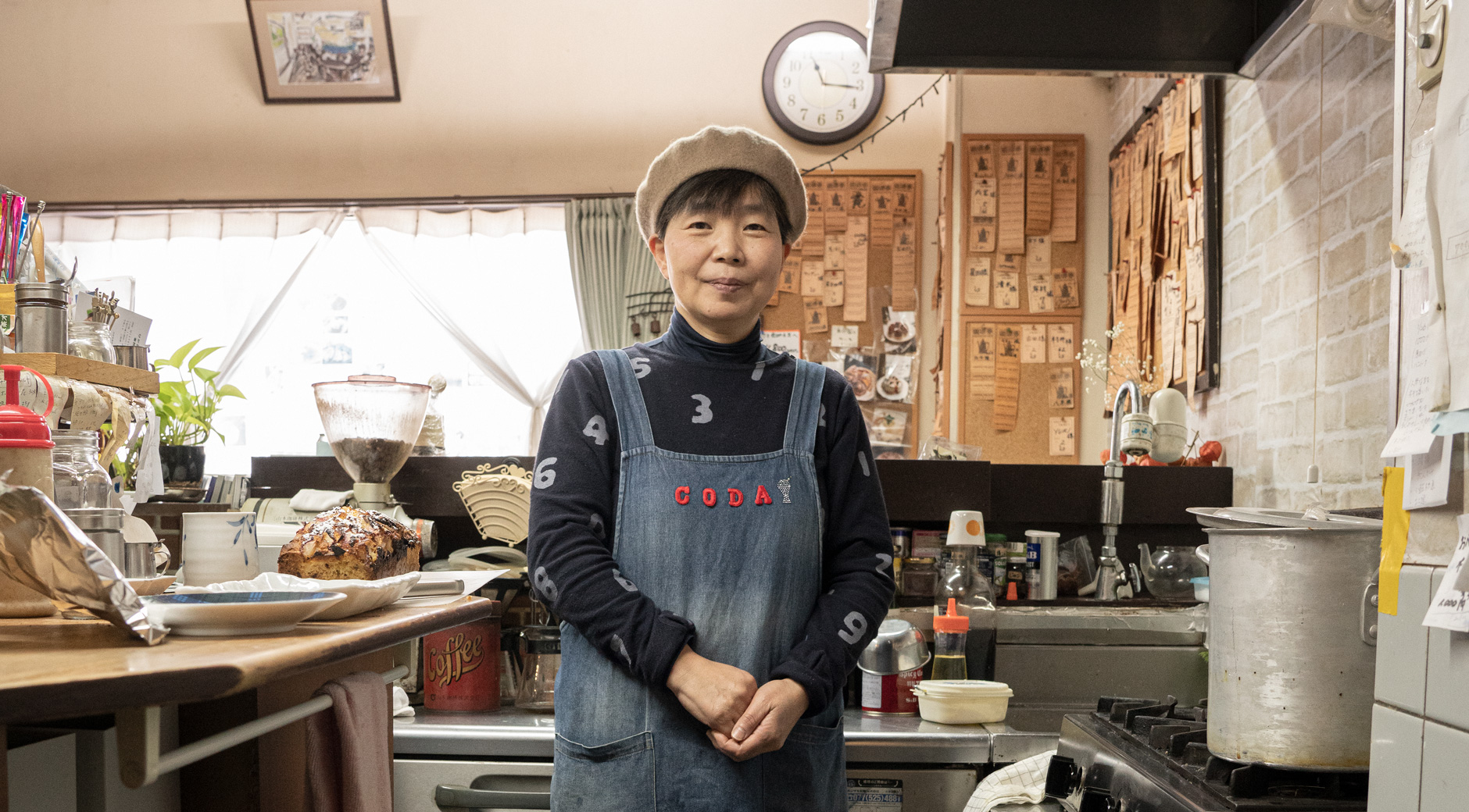 【画像】 カフェの厨房に立つ尾中さん。ショートカットのヘアスタイルにベージュのベレー帽を被り、CODAと赤い文字がアップリケされたデニム生地のエプロンをしている。
