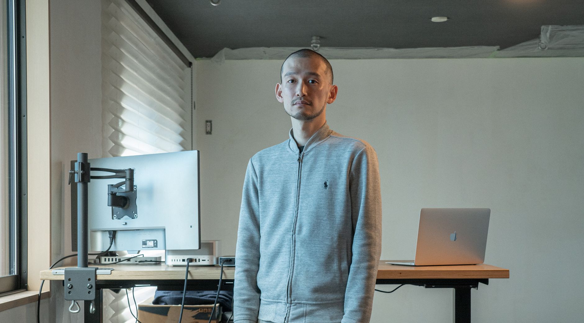 【画像】室内で佇む伊藤浩平さん。髪は刈り上げて短く、グレーのフルジップスウェットにベージュのちのパンツを履いている。伊藤さんの背後にはハイテーブルがあり、ノートパソコンと外付けの大型モニタがある。