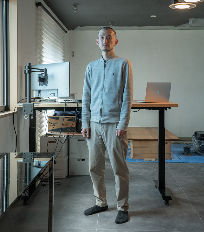 【画像】室内で佇む伊藤浩平さん。髪は刈り上げて短く、グレーのフルジップスウェットにベージュのちのパンツを履いている。伊藤さんの背後にはハイテーブルがあり、ノートパソコンと外付けの大型モニタがある。