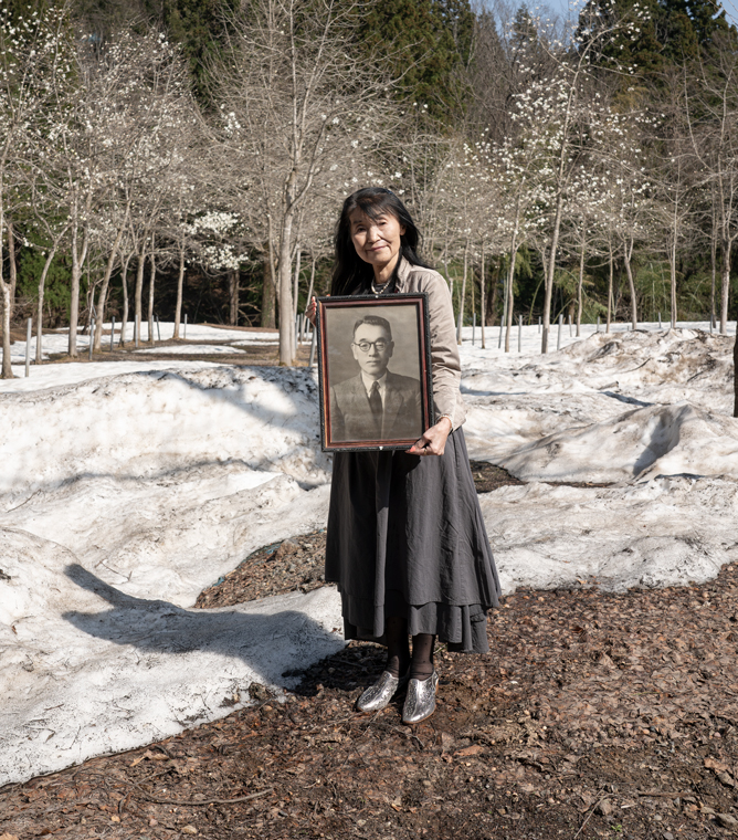 【画像】横尾義智さんのモノクロ肖像写真が入った額縁を手に持ち、屋外に立っている河村一美さん。溶けた雪が地面に広がり、河村さんの背後には冬枯れの樹が並ぶ。