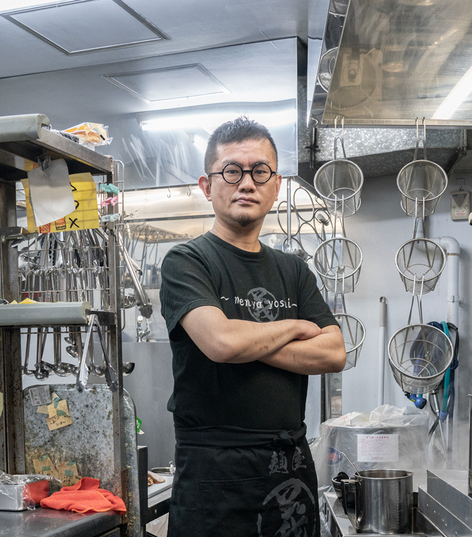 【画像解説】けづかかずよしさんのポートレート写真。ラーメン店の厨房の中で、黒いTシャツに黒い前掛けを着用し、黒縁メガネをかけて腕組みをしている。調理用のトングや網、鍋、コンロなどに囲まれている。