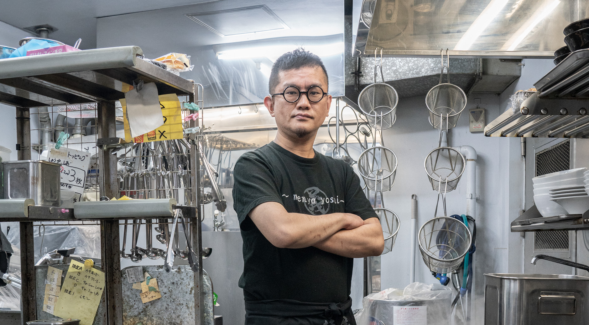 【画像解説】けづかかずよしさんのポートレート写真。ラーメン店の厨房の中で、黒いTシャツに黒い前掛けを着用し、黒縁メガネをかけて腕組みをしている。調理用のトングや網、鍋、コンロなどに囲まれている。