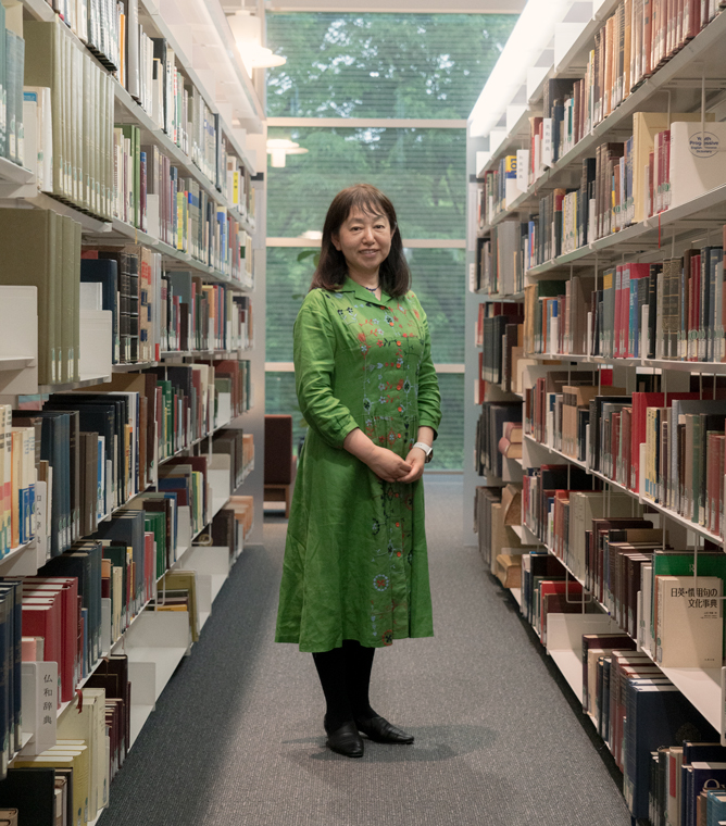 【写真】図書館の中、両側を本棚にはさまれた通路に相良良子さんが立っている。緑色のワンピースを気て、髪は肩までの長さ。両手を前で揃えてこちらを見ている。