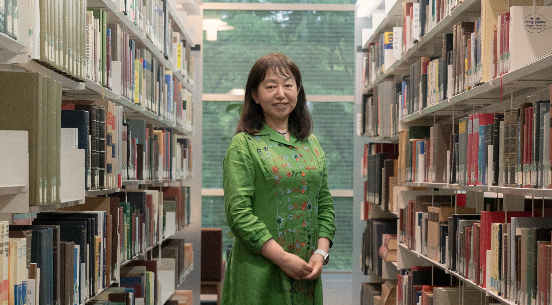 【写真】図書館の中、両側を本棚にはさまれた通路に相良良子さんが立っている。緑色のワンピースを気て、髪は肩までの長さ。両手を前で揃えてこちらを見ている。