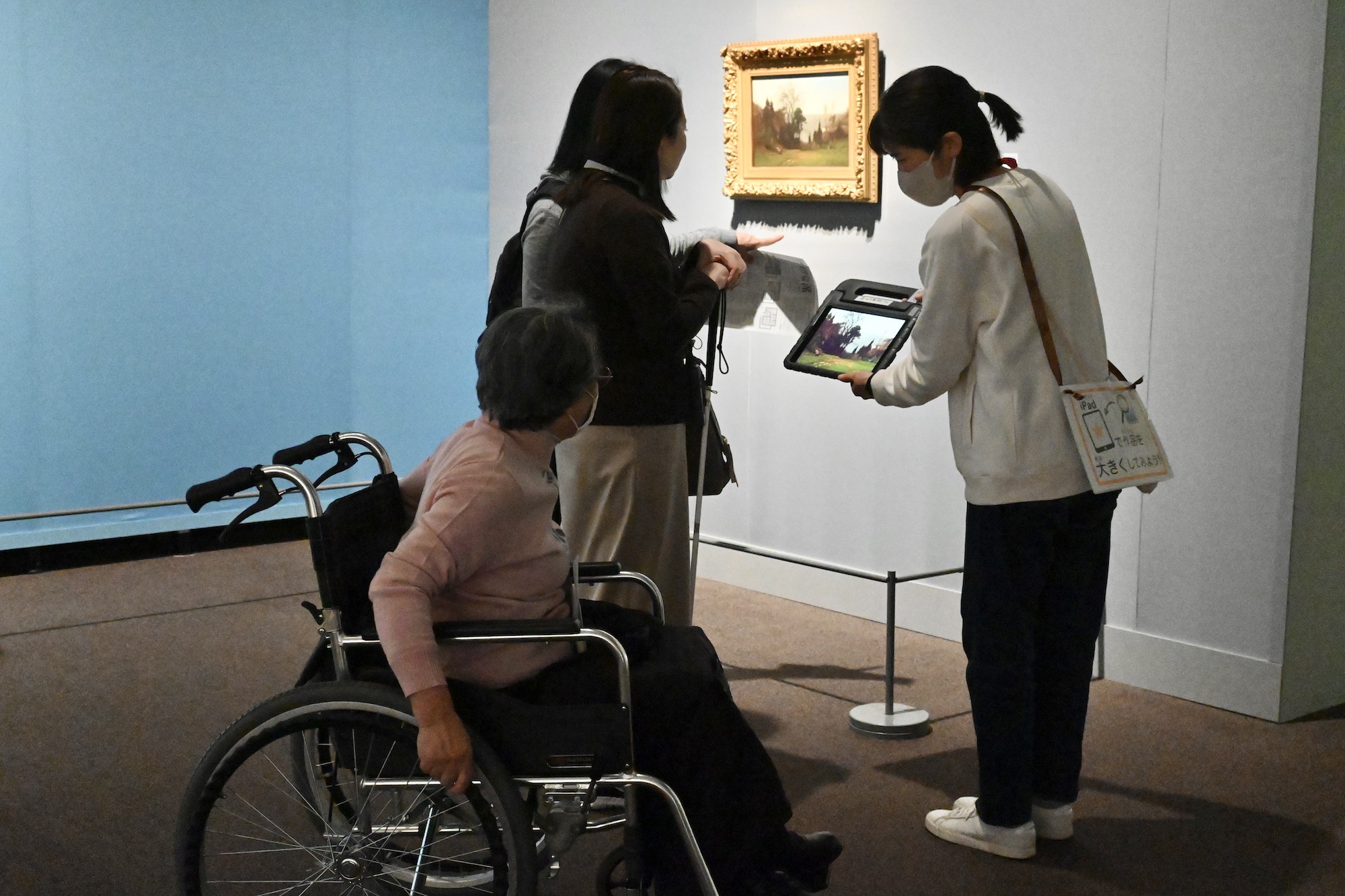 【写真】展示作品の前でタブレット端末を示す女性と、作品を鑑賞する白杖を持った女性と車いすの女性