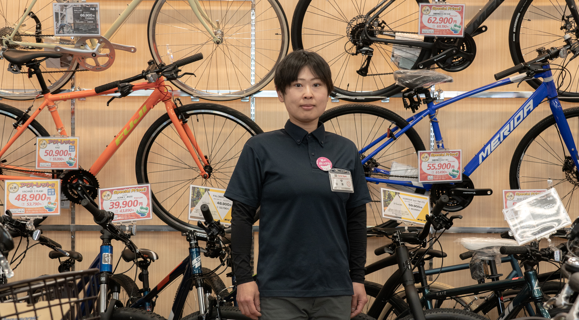 【写真】黒い半袖ポロシャツの下に黒い長袖、グレーのハーフパンツの下に黒いスパッツを着ているショーットカットヘアの鮎澤さん。左胸に名札とピンク色の缶バッチをつけている。後ろにはたくさんのスポーツ自転車が並んでいる