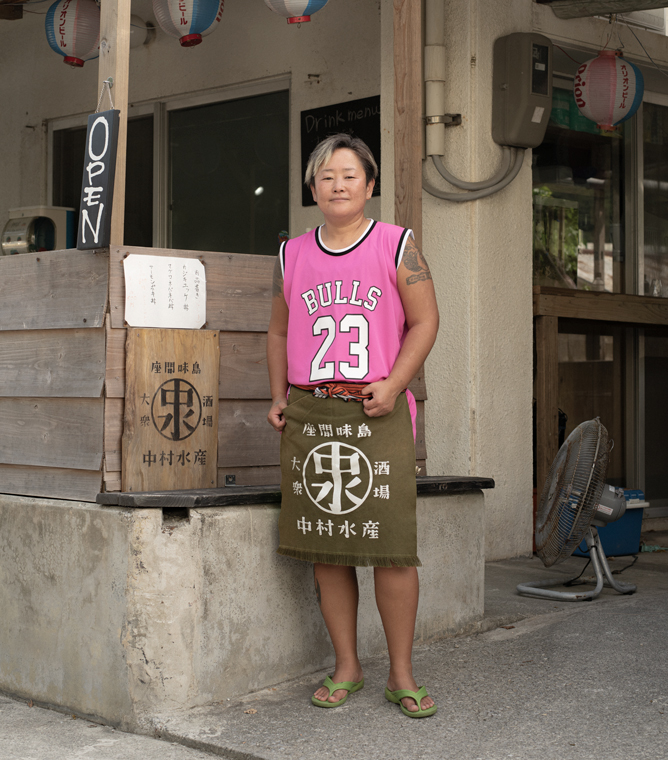 【写真】お店の前で中村水産の前掛けをかけて立っているチアキさん。日に焼けた肌でショートカット、前髪にはメッシュ、鮮やかなピンク色のバスケットユニフォームと黄緑色のビーチサンダルを履いている