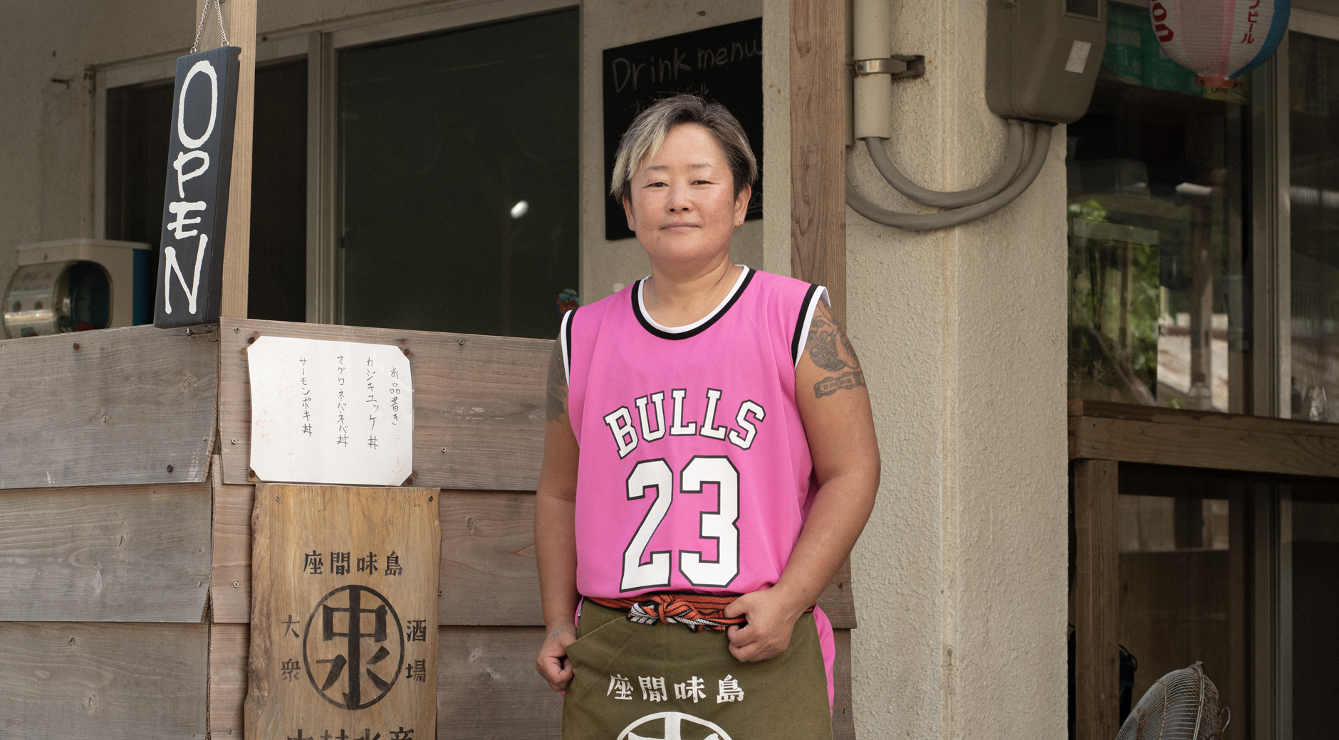 【写真】お店の前で中村水産の前掛けをかけて立っているチアキさん。日に焼けた肌でショートカット、前髪にはメッシュ、鮮やかなピンク色のバスケットユニフォームと黄緑色のビーチサンダルを履いている