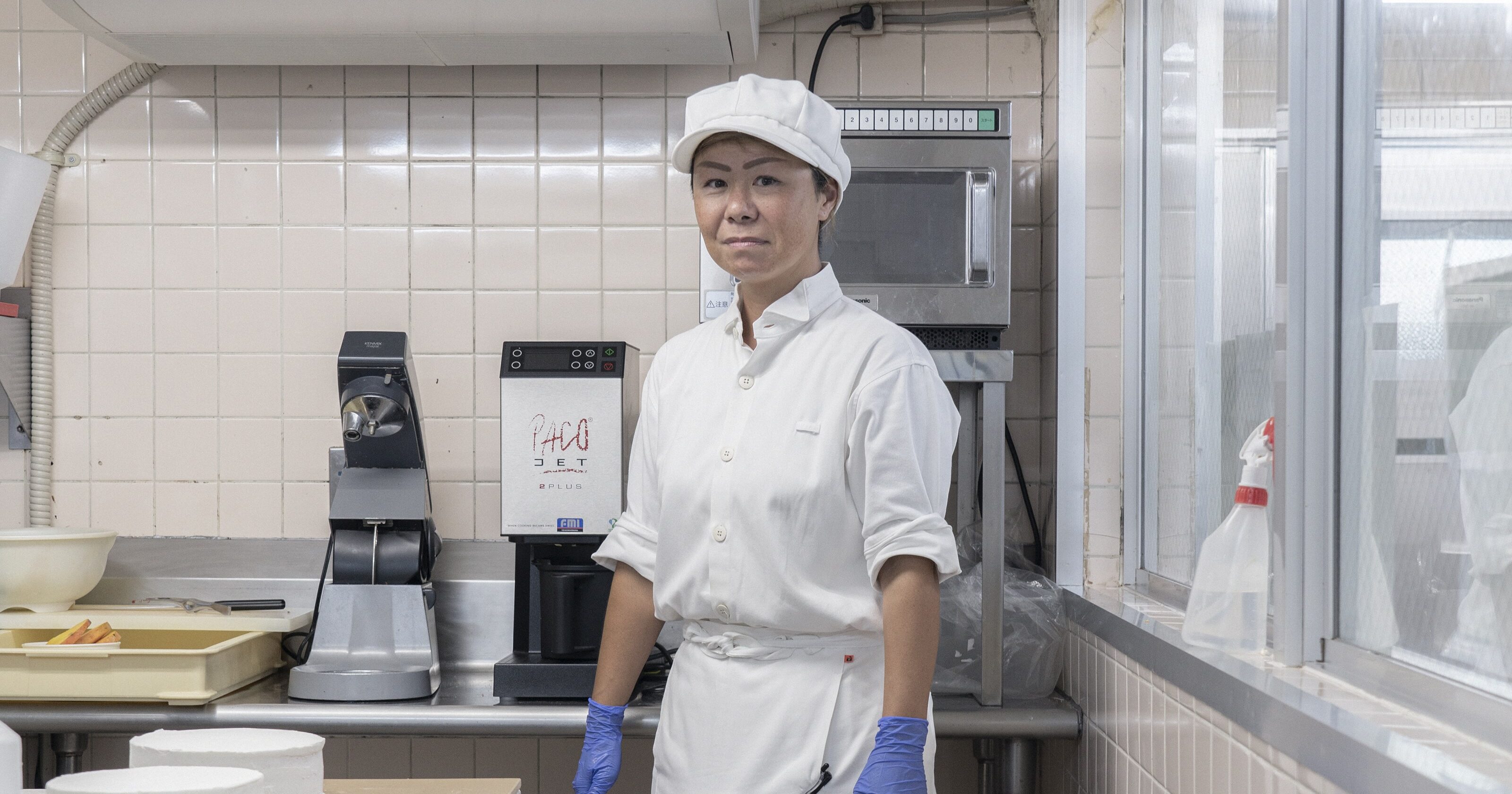 【写真】調理室で、白い帽子、白いシャツ、白いロングエプロンをみにつけて、青いゴム手袋をしている、しまぬきまなみさん。調理台の上にはつくりかけのホールケーキが２つ載っている。