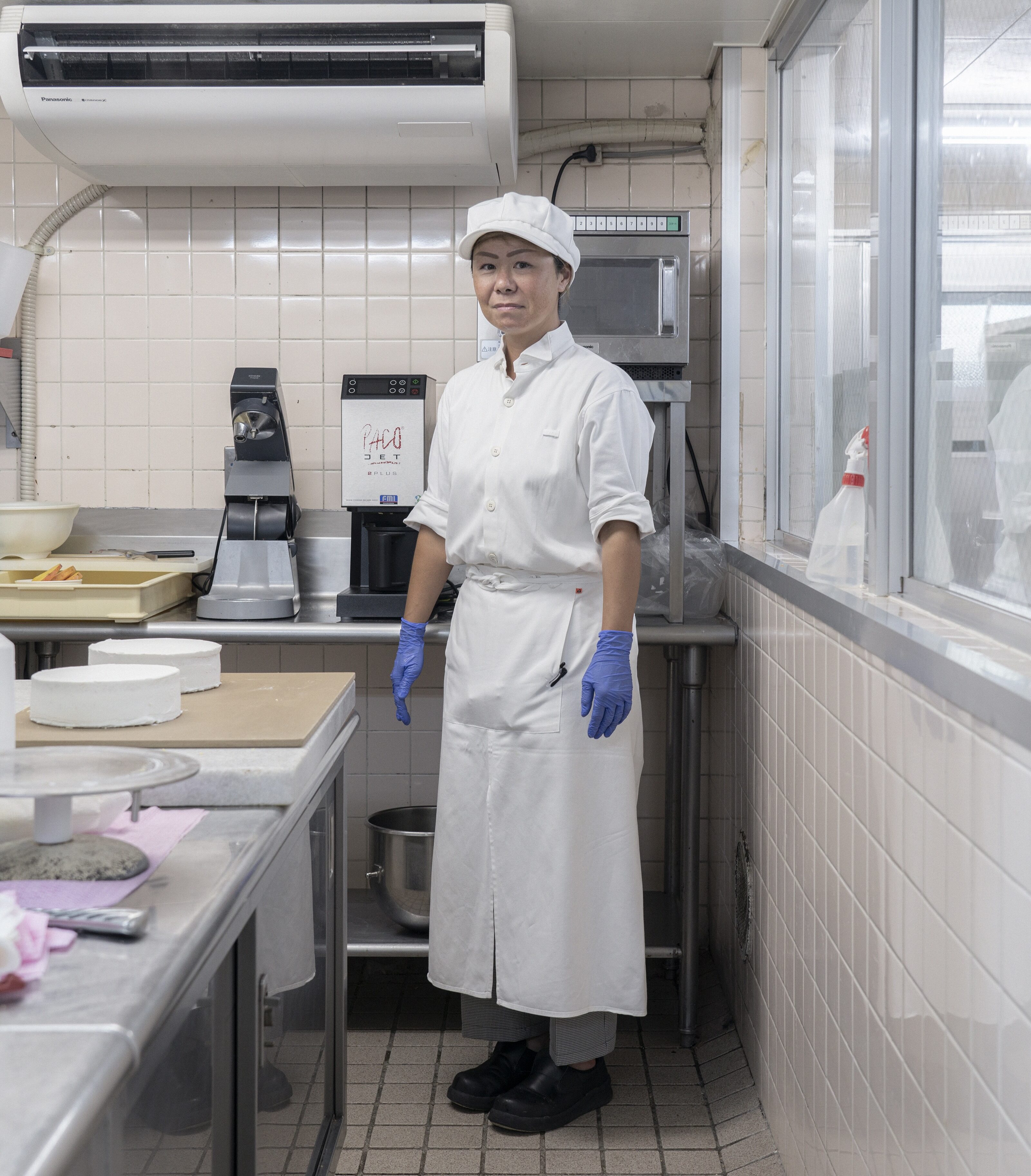 【写真】調理室で、白い帽子、白いシャツ、白いロングエプロンをみにつけて、青いゴム手袋をしている、しまぬきまなみさん。調理台の上にはつくりかけのホールケーキが２つ載っている。