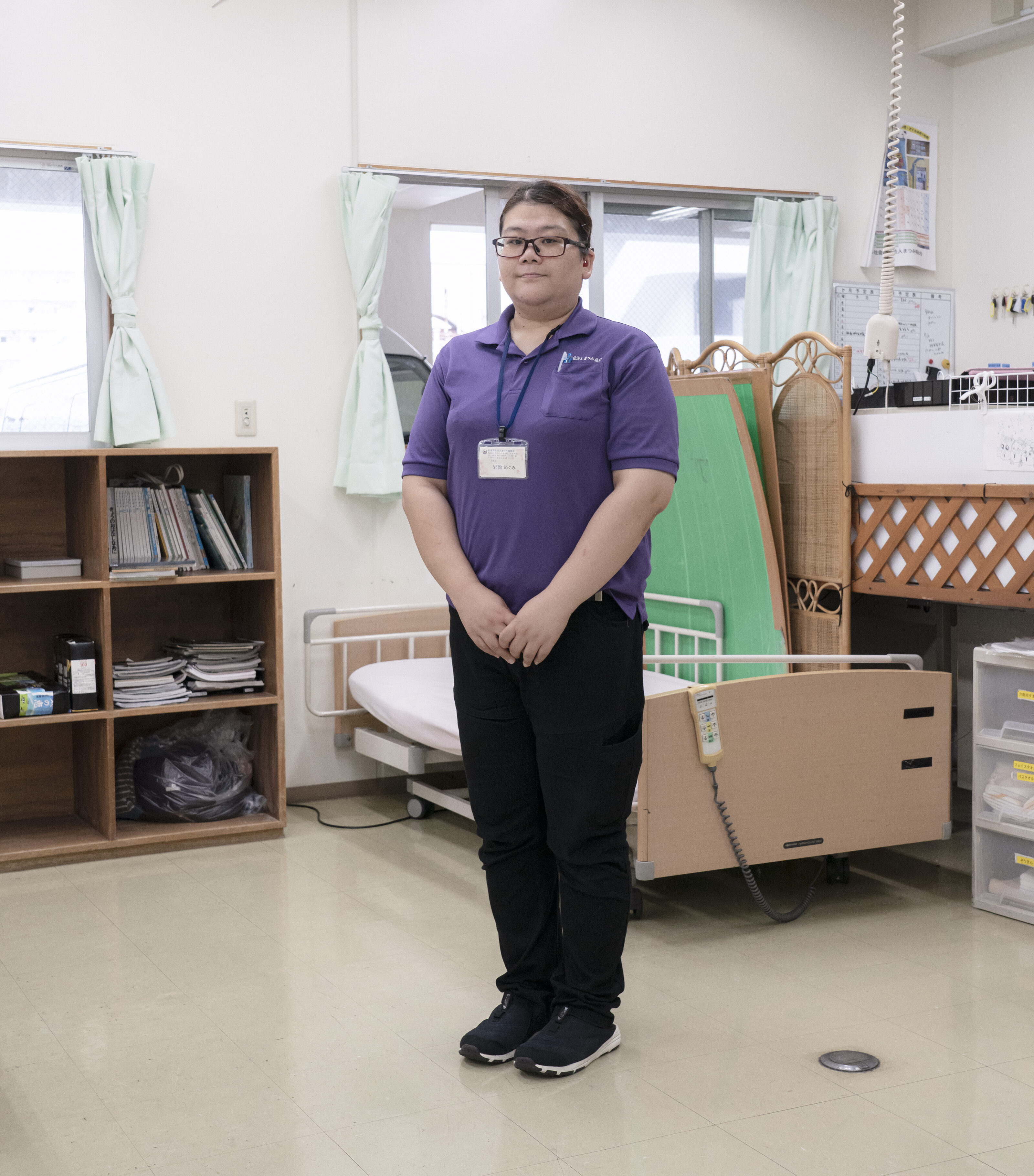 【写真】介護用ベッドのそばに立ち、こちらを眺めるいわだてさん。紫のポロシャツを着て、メガネとスタッフ証をかけている。