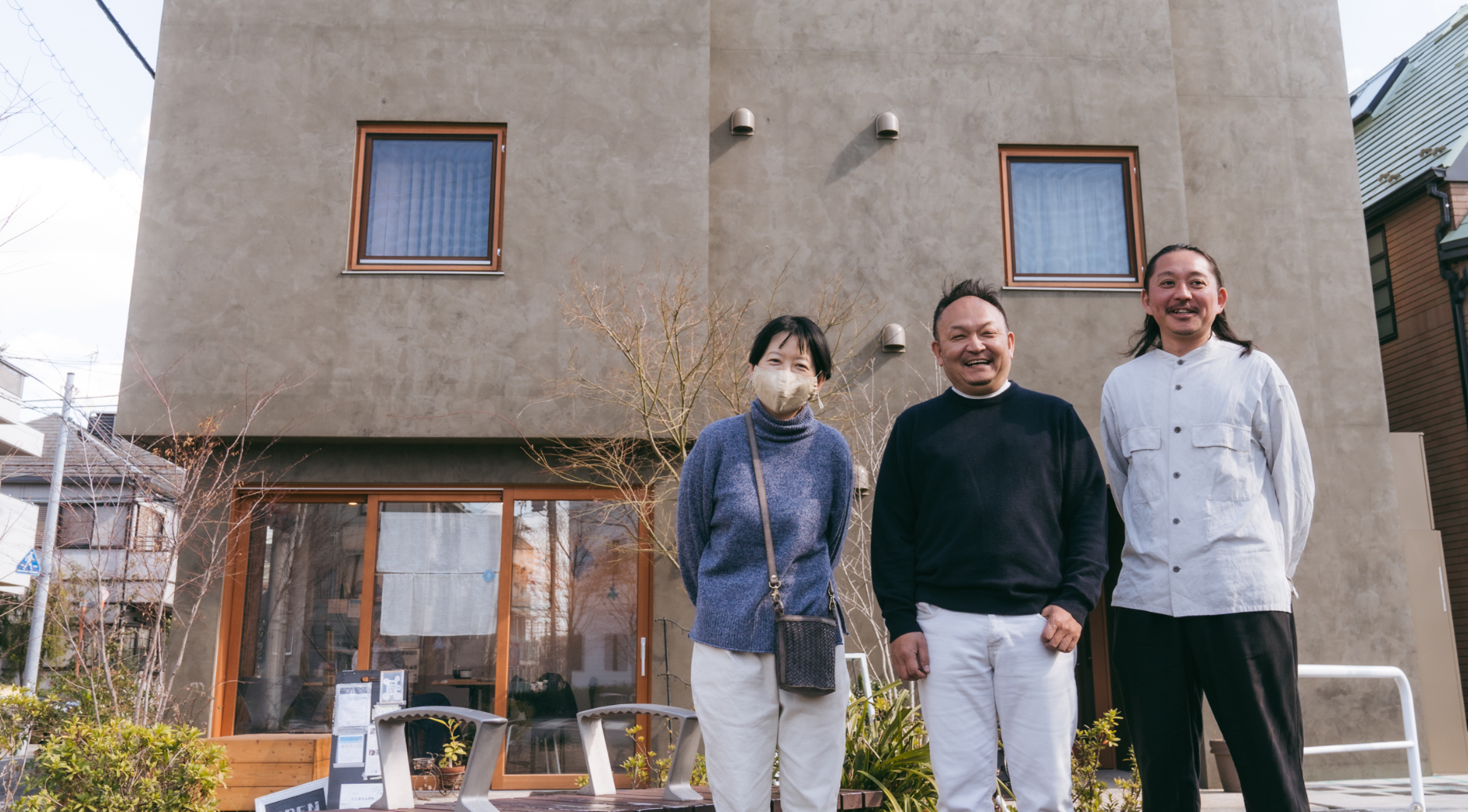 【写真】薄茶色の壁、木の枠の大きな窓が印象的な建物の前で3人の男女が並んでいる