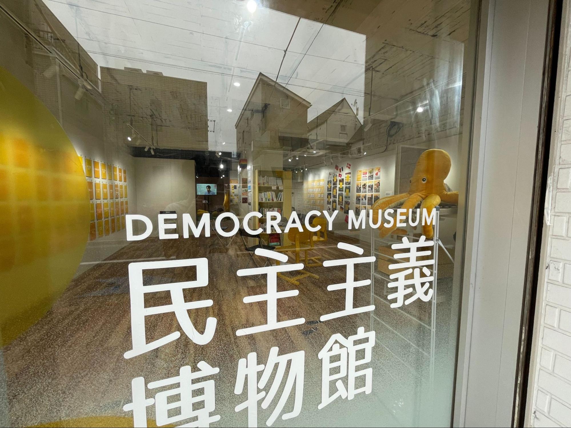 【写真】民主主義博物館の文字が入った大きなガラスウィンドウ。中では、左右の壁を中心にさまざまな展示があることがわかる