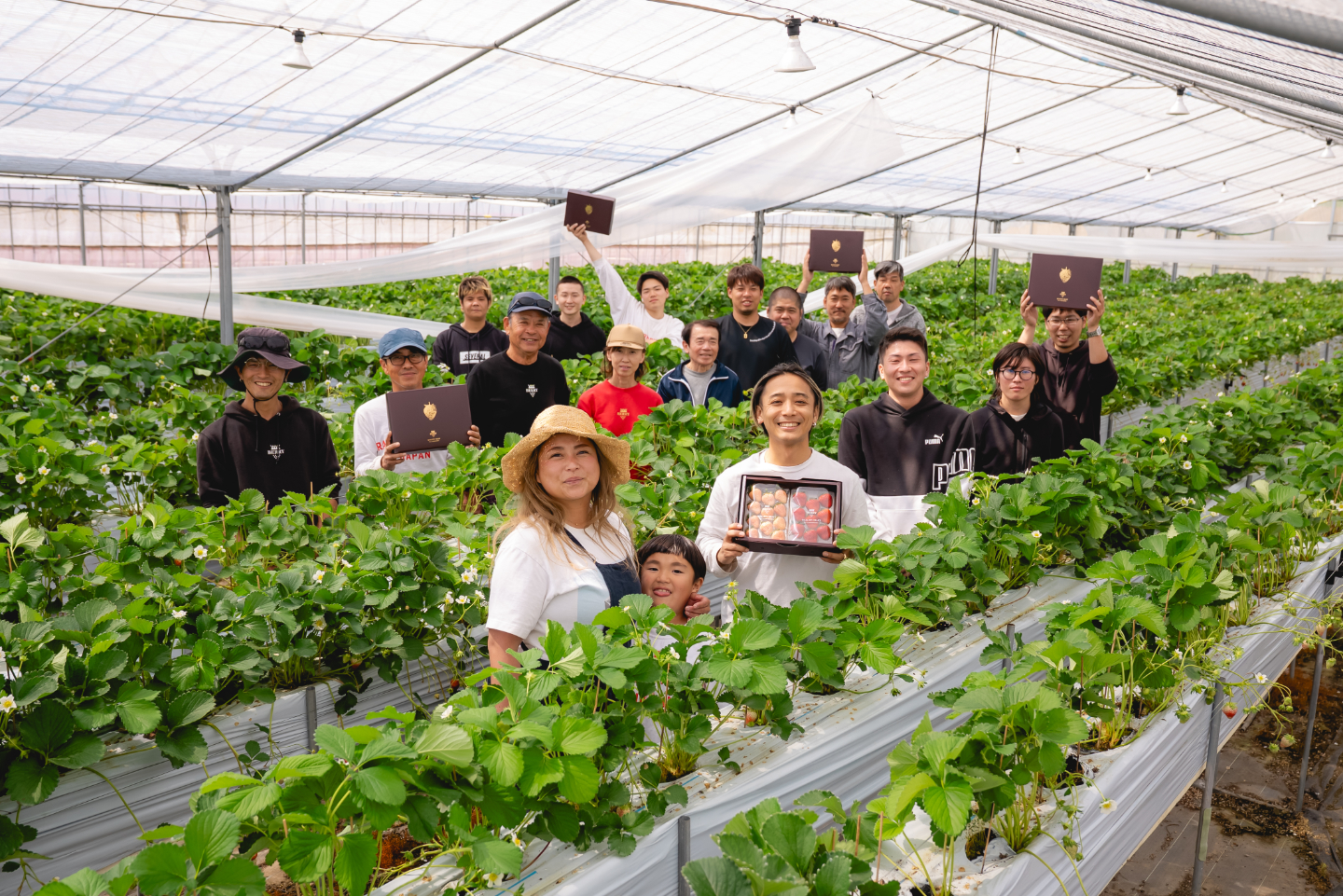 【写真】ハウスのなかで、たくさんの人がこちらを向いている。中央に、いちごが入った箱を笑顔で掲げる宮澤さん