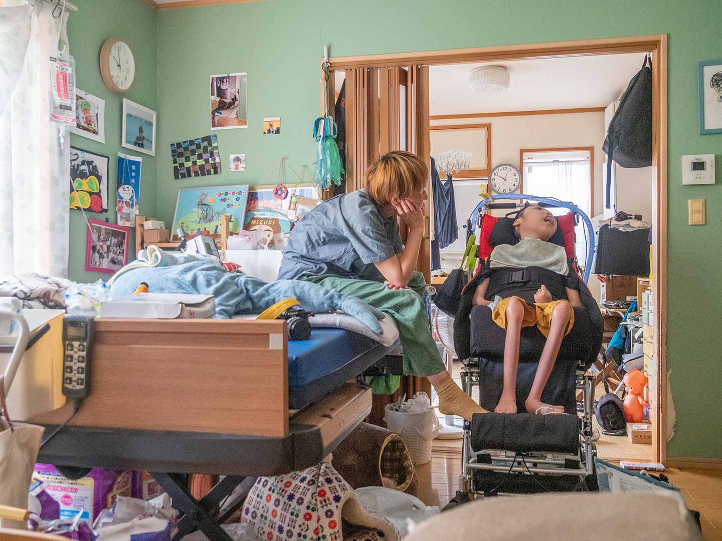 山本さんのご自宅の息子さんの部屋の写真。車椅子に乗る息子さんと、ベッドに座り息子さんを眺める山本さんを捉えた写真。部屋の様子は、若葉色の壁紙に、さまざまな写真やイラストなどが貼り出されてある。