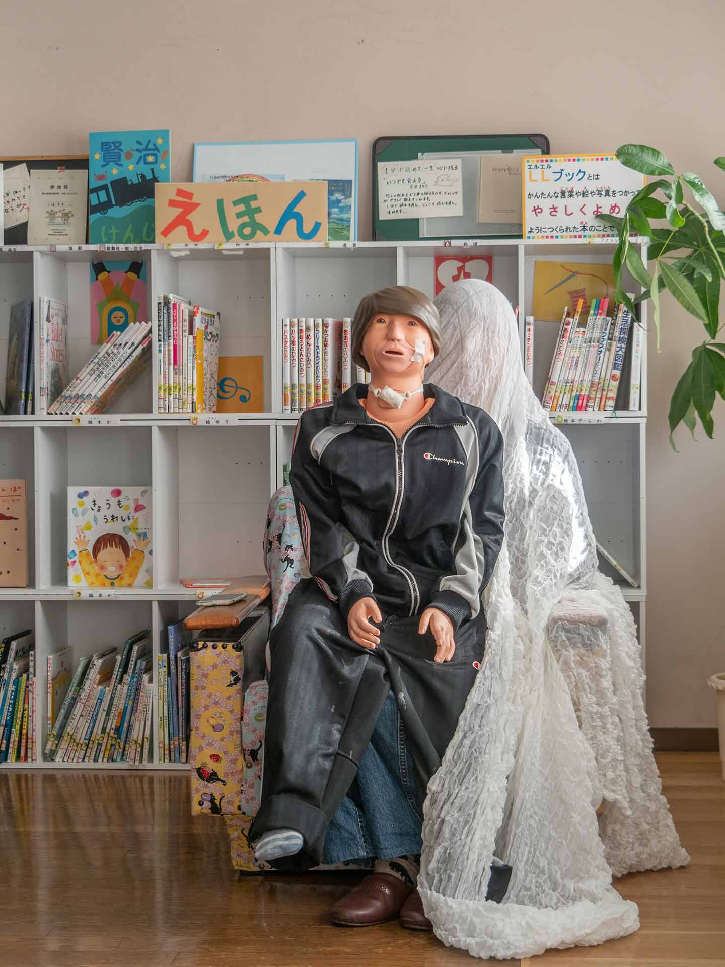 さまざまな絵本が置かれた学校内の本棚の前で、白いヴェールを全身に被り、小児への基本看護技術の習得や病児の日常的なサポート技術のトレーニングに使用する小児看護実習モデルの「まぁちゃん」を背後から抱える山本さんのポートレート。