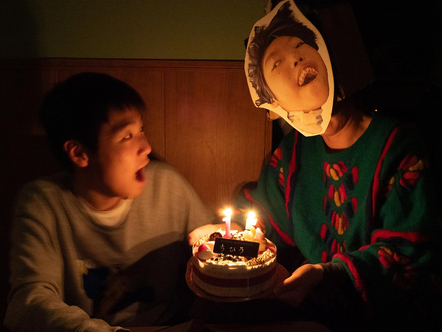 亡くなった医療的ケア児だった息子さんの顔写真を切り取ったお面を被り、火のついたバースデーケーキを抱え、別の息子さんの誕生日を祝う山本さんの写真。