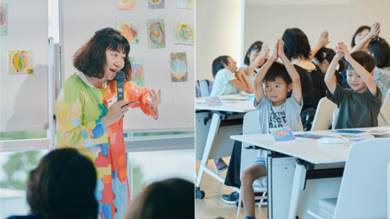 【写真】ホワイトボードに貼られたたくさんの絵の前で話す人と、手を三角にして頭の上にあげるたくさんの子どもたち