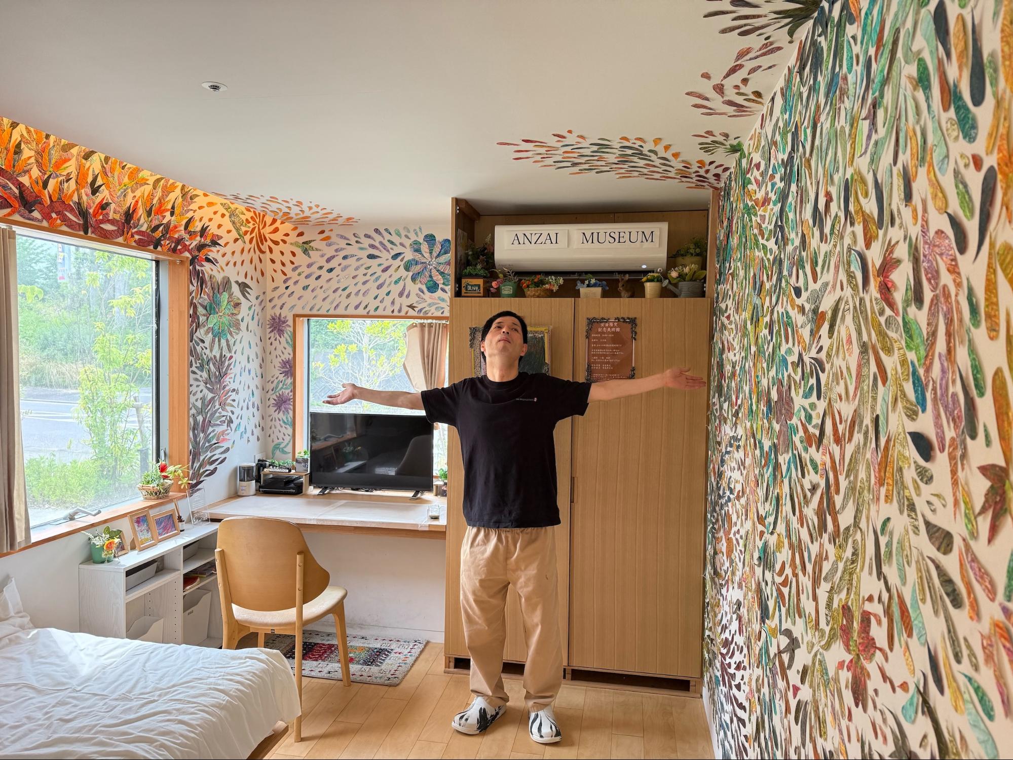【写真】壁にカラフルなイラストが一面描かれた、ホテルの一室のような部屋に男性がたたずむ