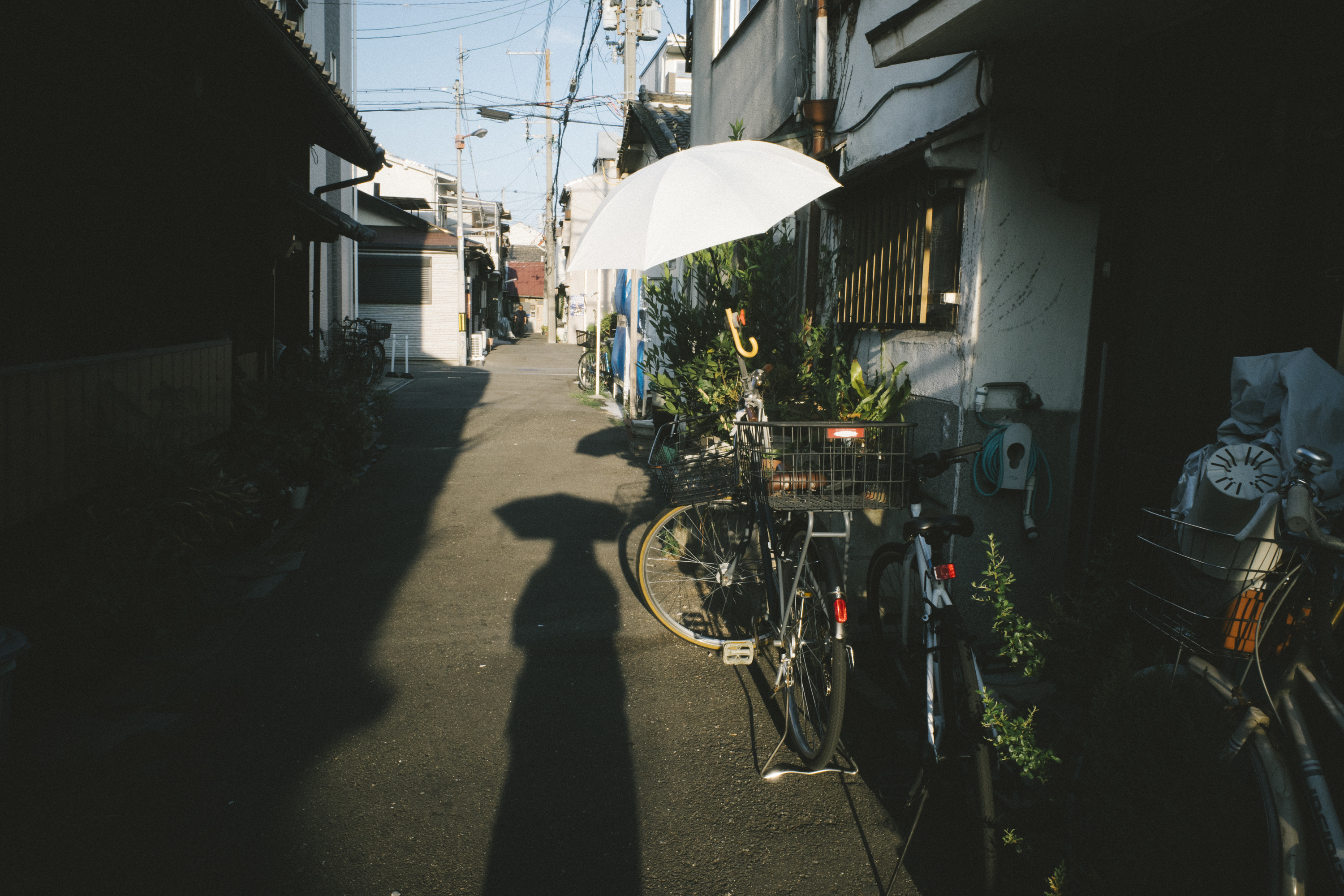 【写真】住宅街、コンクリートで舗装された道の上に自転車が止まっている。その自転車には傘。地面には、それを眺めているであろう人の影が出ている。