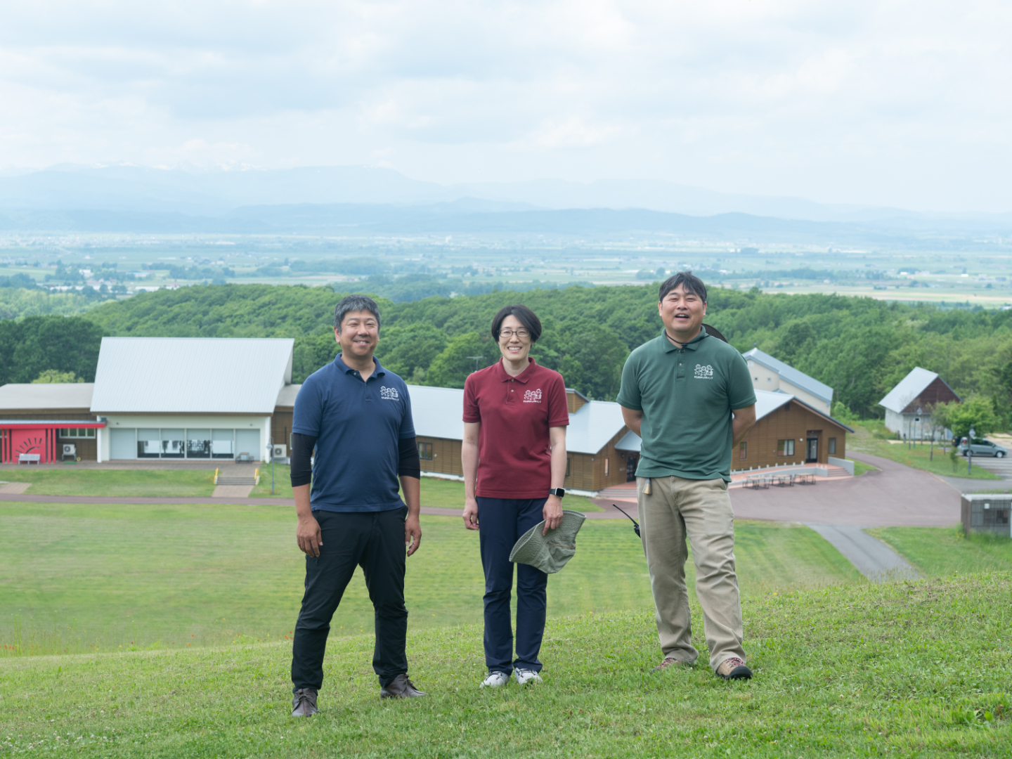 【写真】丘の上に立ちこちらを向く3人。背景には青空と芝生、複数の施設