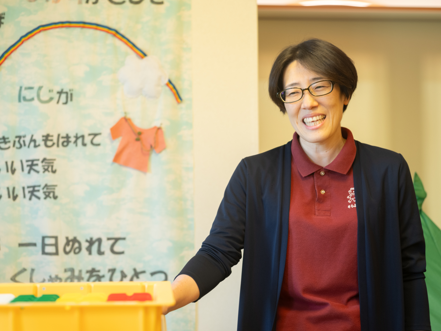 【写真】にじ、の歌が書かれた壁面の横で、笑顔で立つ宮坂さん