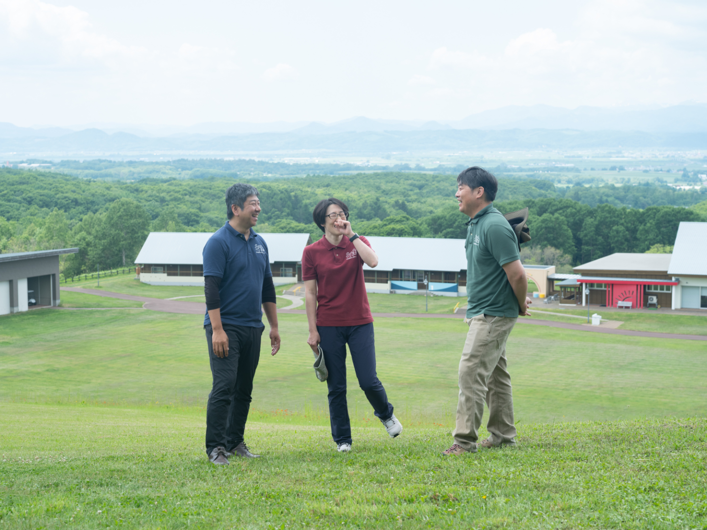【写真】芝生の丘の上で、3人が楽しそうに会話をしている。背景には複数の建物と広がる森、遠くには山並み