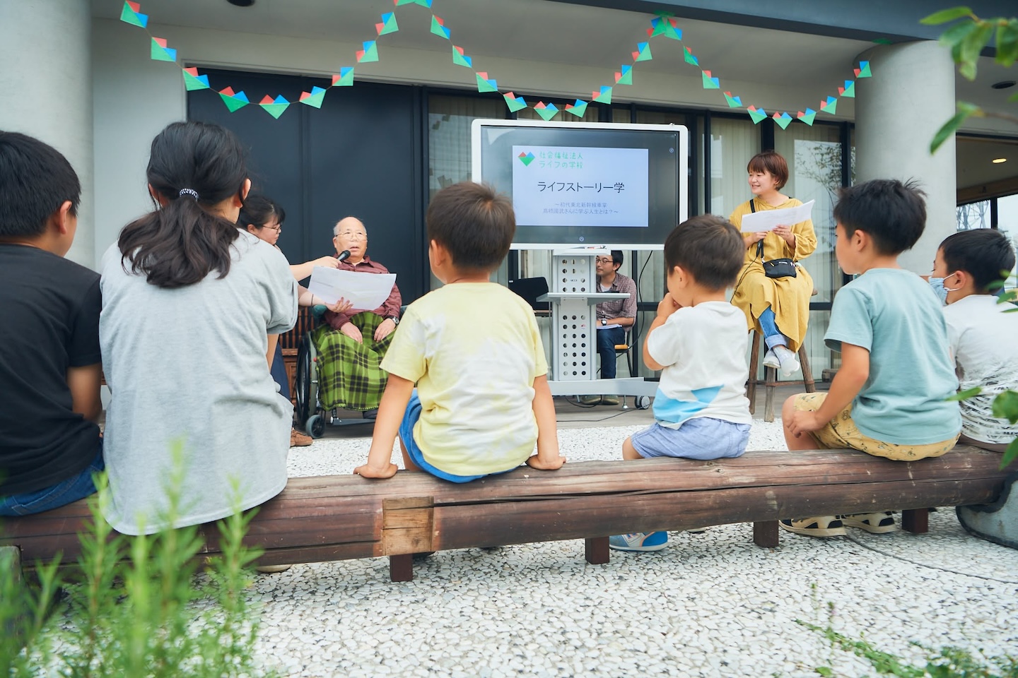 奥にマイクを向けられたパートナーが座り、隣にはスライドが映っている手前のベンチには子どもたちが座って、話を聞いている