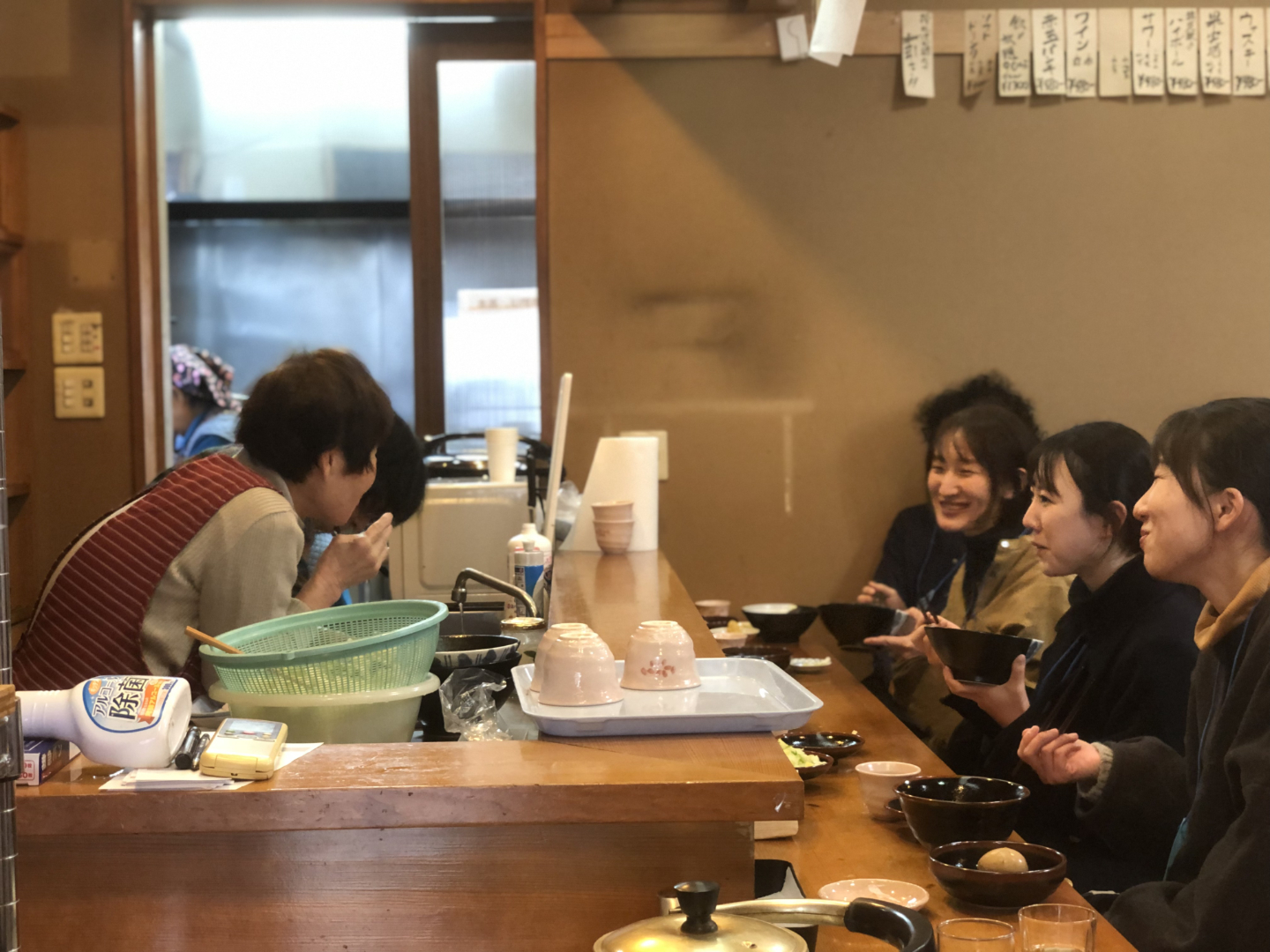 左側のキッチンの中で話している様子のおばあちゃんに、右側のカウンターに並んで食事をしている人たちが笑っている