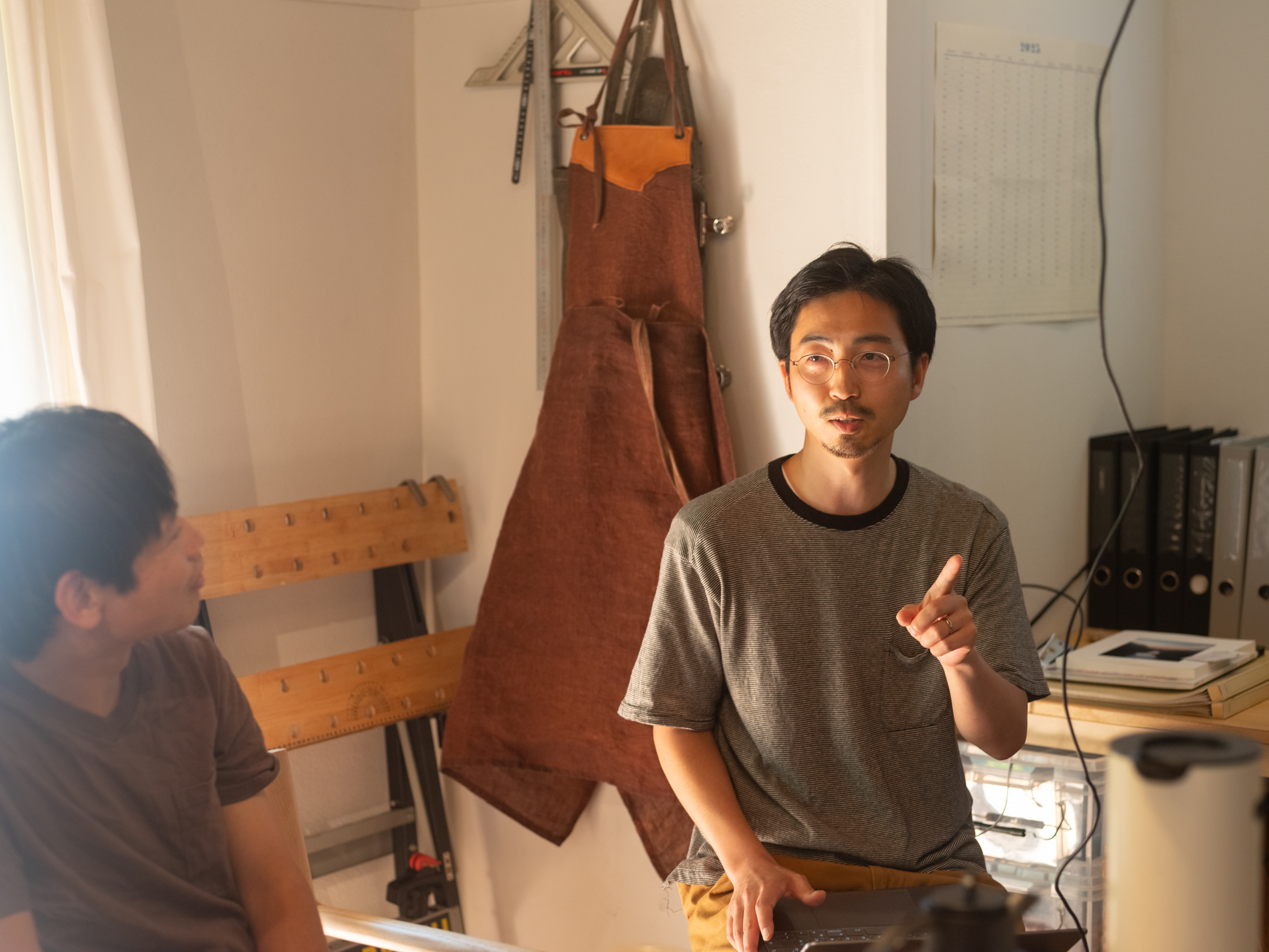 【写真】二人の人物が写真にうつっている。左側の人物は右側の人物をみていて、右側の人物は短髪にメガネ、正面をみながら左手の人差し指をたてている。