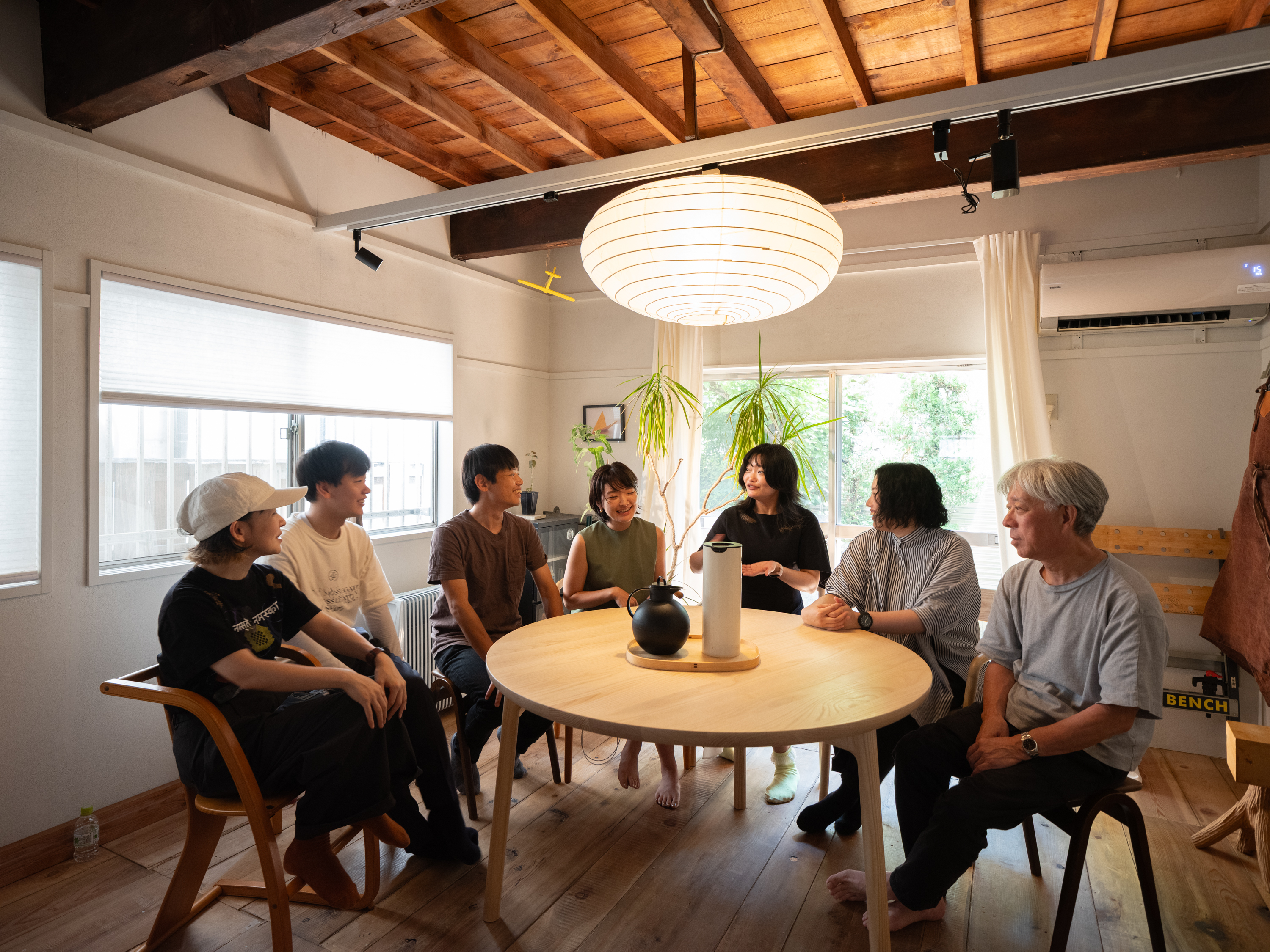 【写真】丸いダイニングテーブルの向こう側に〈こここ〉編集部の7名が座っている。テーブルの上にはポットが２つ並び、天井からは和紙でできた平たい円形の照明がさがっている。メンバーは歓談している。