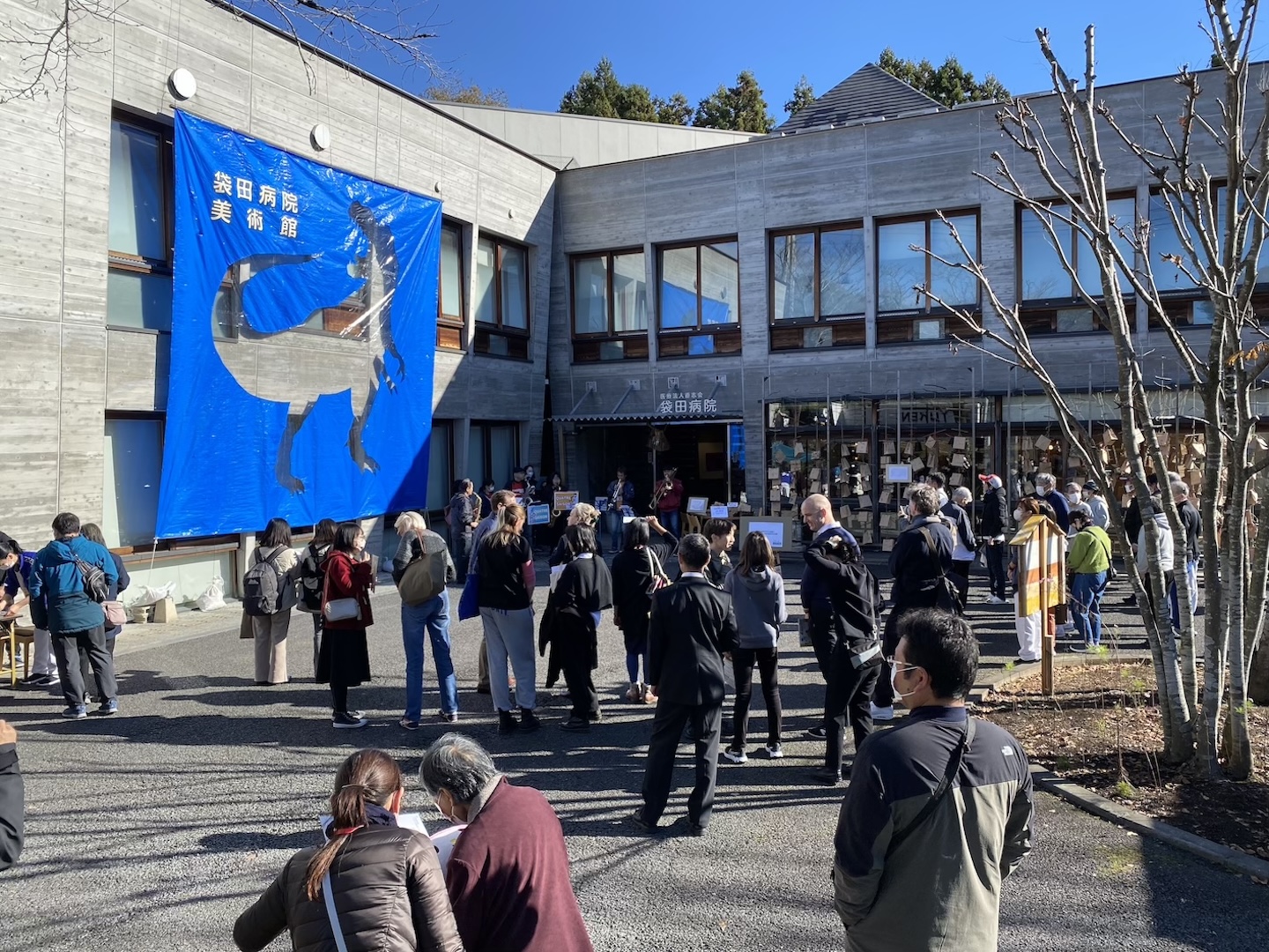 病院の建物の外側にたくさんの人。建物には「袋田病院美術館」と書かれた、恐竜の絵がくり抜かれた大きなポスターが飾られている