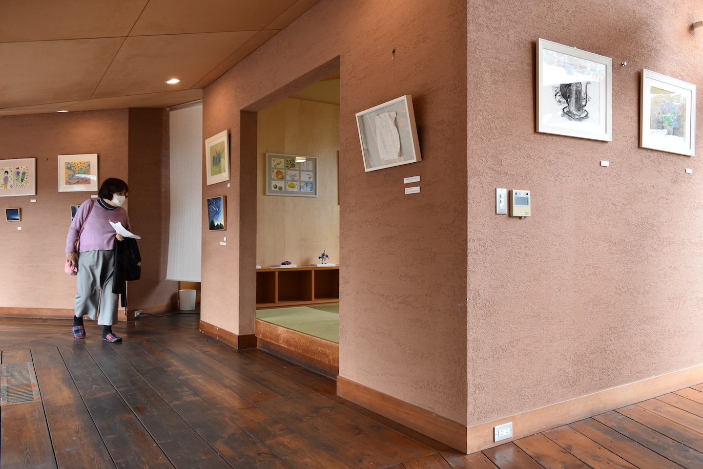 ピンク色の壁に絵が飾られ、歩きながら見ている人がいる