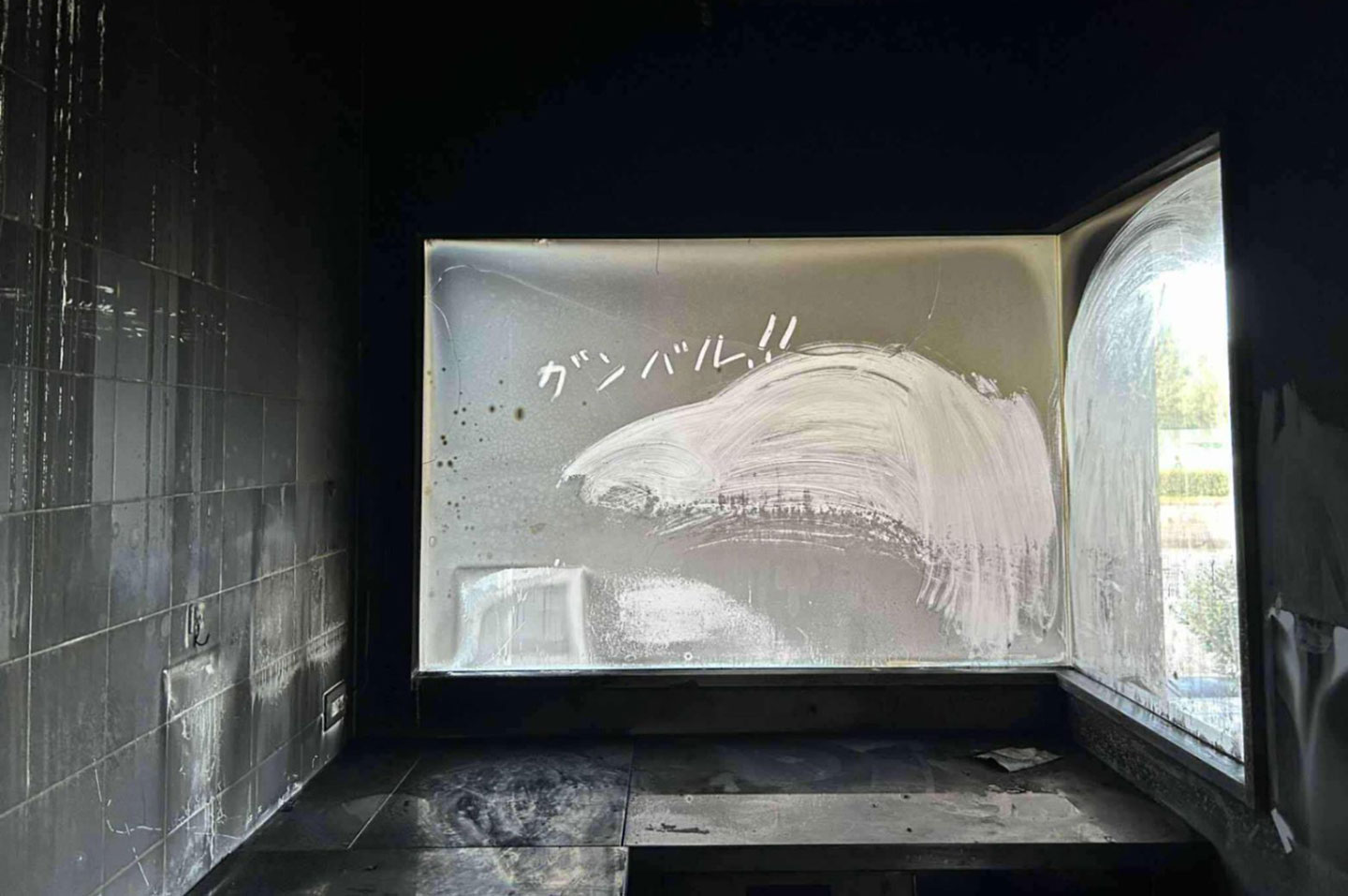 「ガンバル!!」と煤だらけのガラス窓に書かれた厨房の写真。