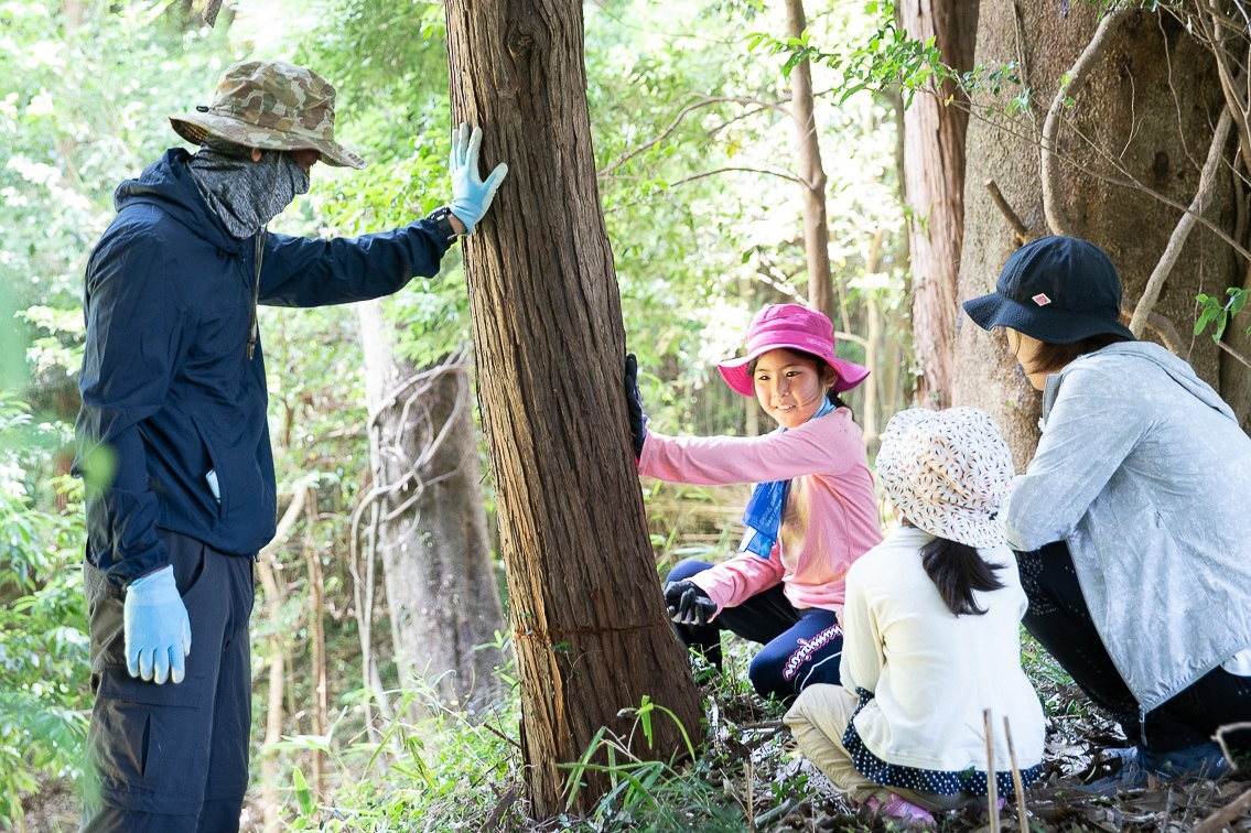 【写真】森にある木に手を置いて話す人、横に佇む親子