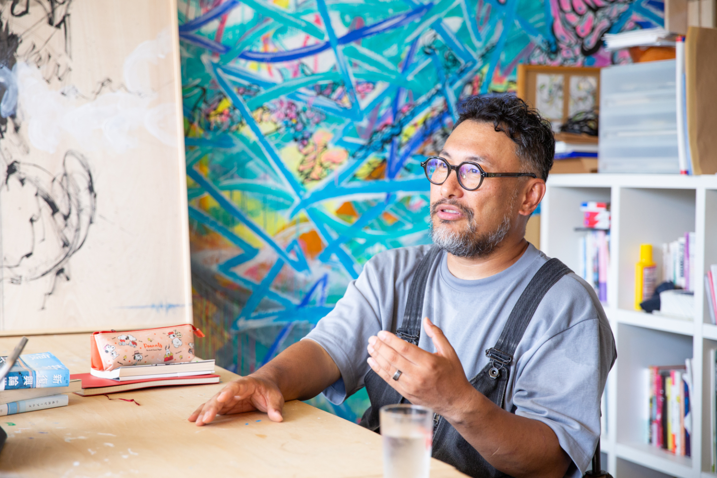 【写真】テーブルに座って話す小松さんの横顔。背景の壁には大きな壁画