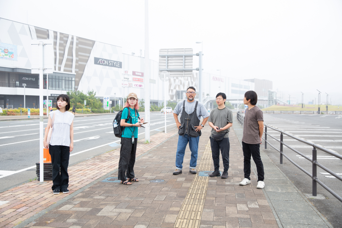 【写真】片側が広い道路、片側が駐車場になっている間の歩道で話をする5人。中央に小松さん、青山さん