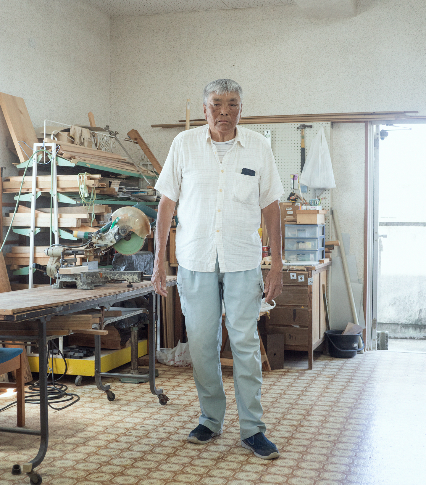 【写真】室内で木材が積まれ棚の前に立つ、たなかみちおさん。白髪、日に焼けた肌、白い半袖シャツ、ブルージンズ。