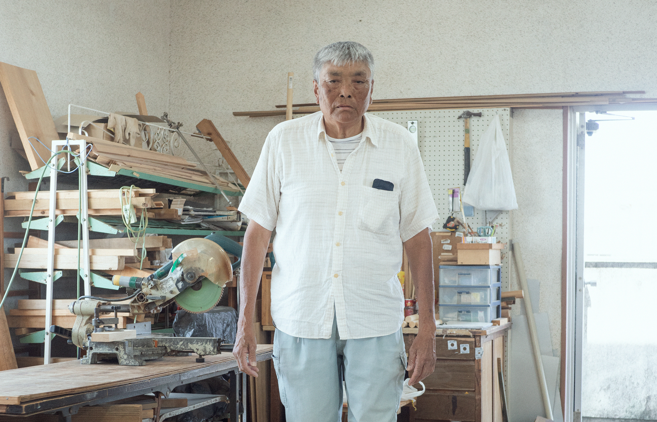 【写真】室内で木材が積まれ棚の前に立つ、たなかみちおさん。白髪、日に焼けた肌、白い半袖シャツ、ブルージンズ。