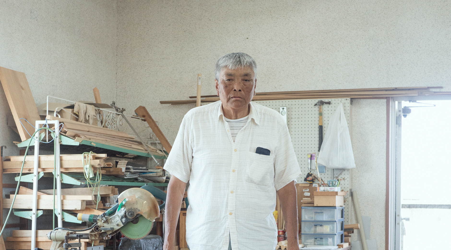 【写真】室内で木材が積まれ棚の前に立つ、たなかみちおさん。白髪、日に焼けた肌、白い半袖シャツ、ブルージンズ。
