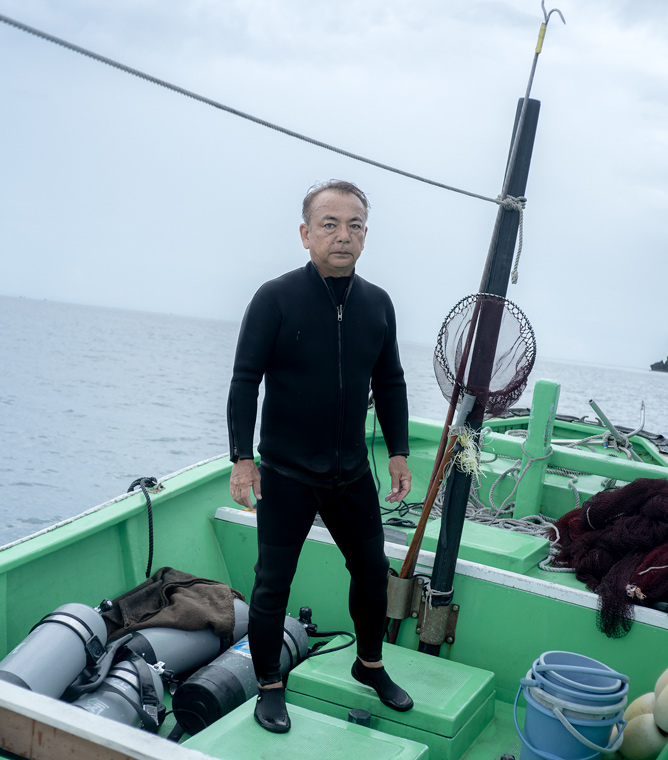 【写真】海の上に浮かぶ船に立つ山下さん。黒いウェットスーツの上下を着て、両手を身体の横に下げる形で正面を見つめている。船の上にはタモやバケツ、空気ボンベなどの道具が並んでいる。
