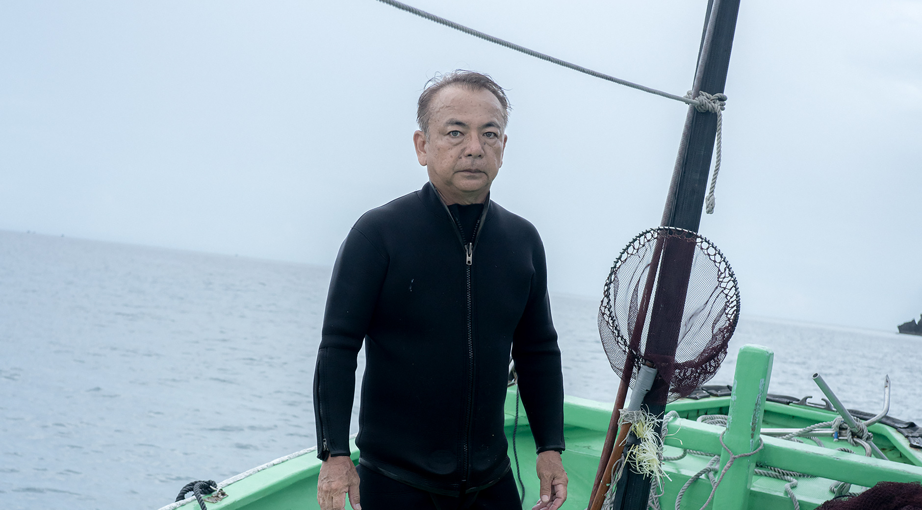 【写真】海の上に浮かぶ船に立つ山下さん。黒いウェットスーツの上下を着て、両手を身体の横に下げる形で正面を見つめている。船の上にはタモやバケツ、空気ボンベなどの道具が並んでいる。