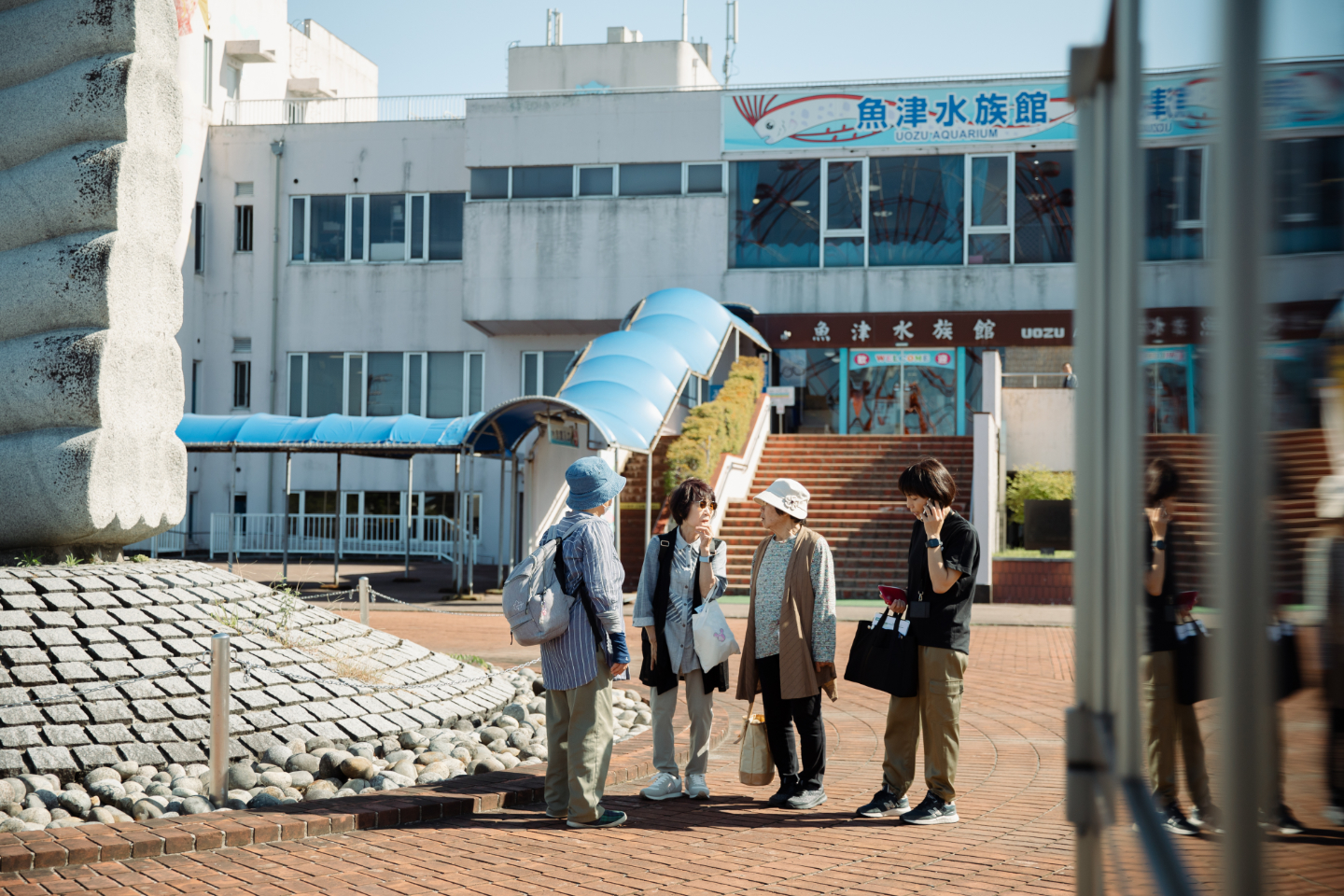 【写真】晴れた日に魚津水族館の入口前で高齢者のグループが立ち話をしている。帽子やかばんを持ち外出中であることが伝わる