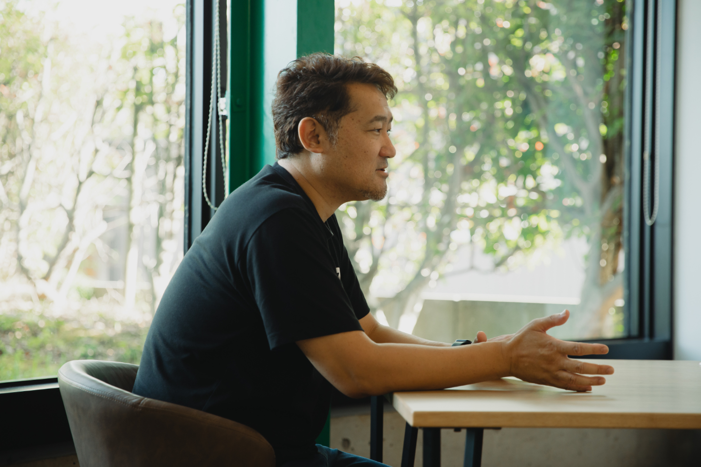【写真】室内のテーブルで小柴さんが身振りを交えて話している。窓の外には緑が見える