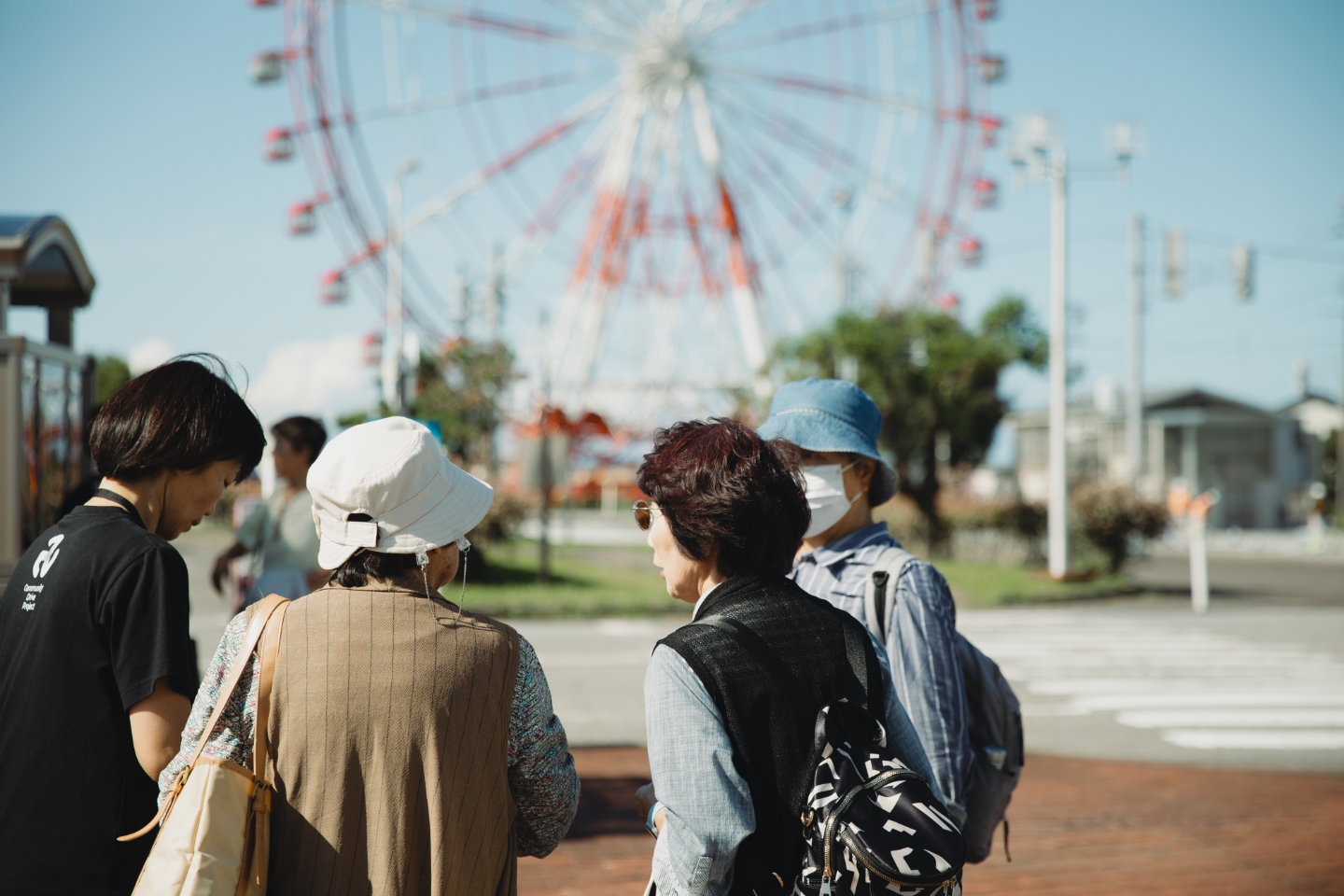 【写真】屋外で高齢者とスタッフが集まり会話している後ろ姿。背景には大きな観覧車が見える